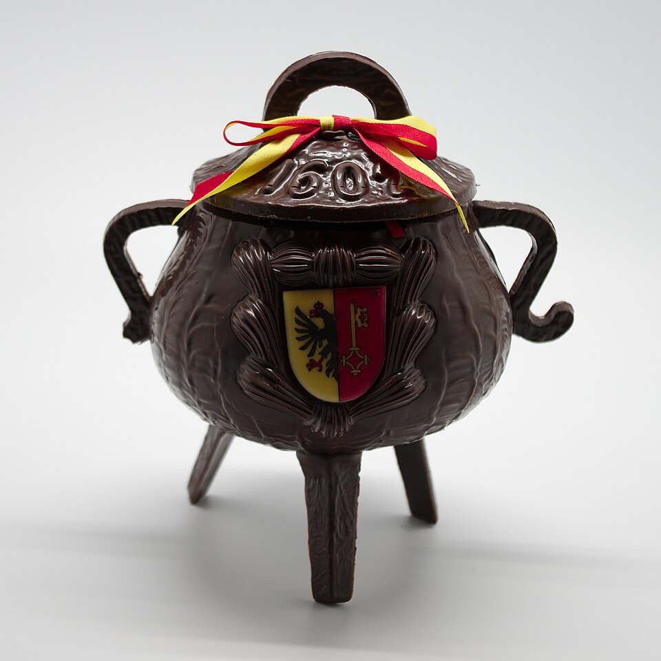 Photographie illustrative : Marmite de l’Escalade en chocolat ornée des couleurs genevoises, photographiée sur fond clair.