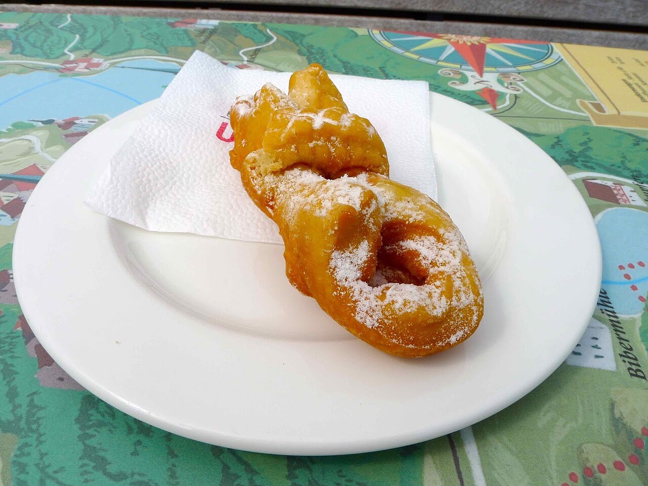 Photographie illustrative : Deux beignets dorés et torsadés de Schleitheim, saupoudrés de sucre glace, sur une assiette blanche.